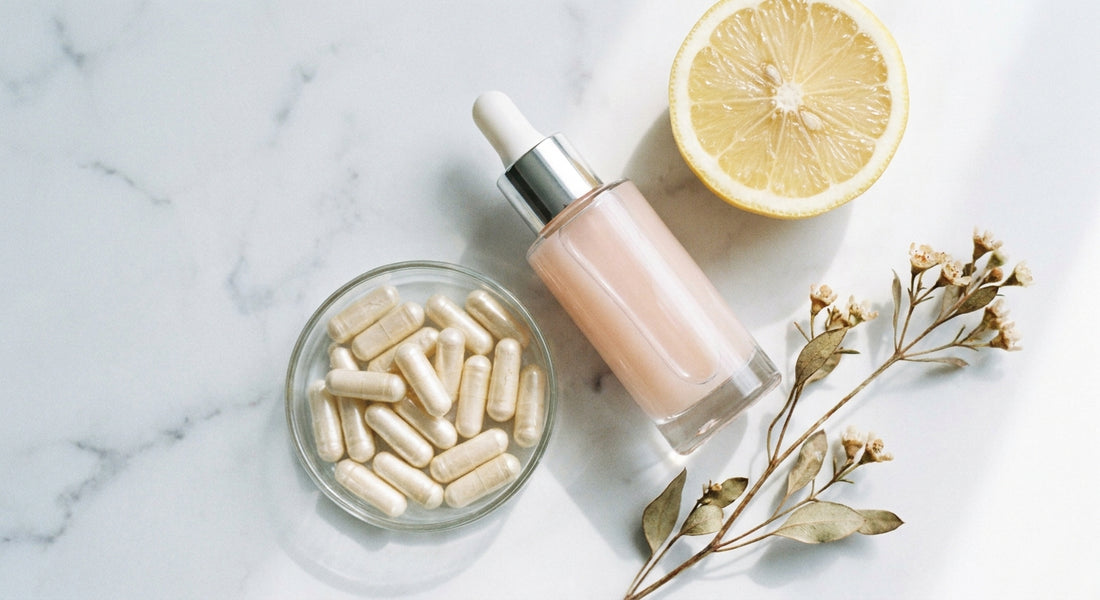 A minimalist marble flat-lay of a glass serum dropper bottle alongside supplement capsules, a sliced citrus fruit, and botanical sprigs in soft natural light.