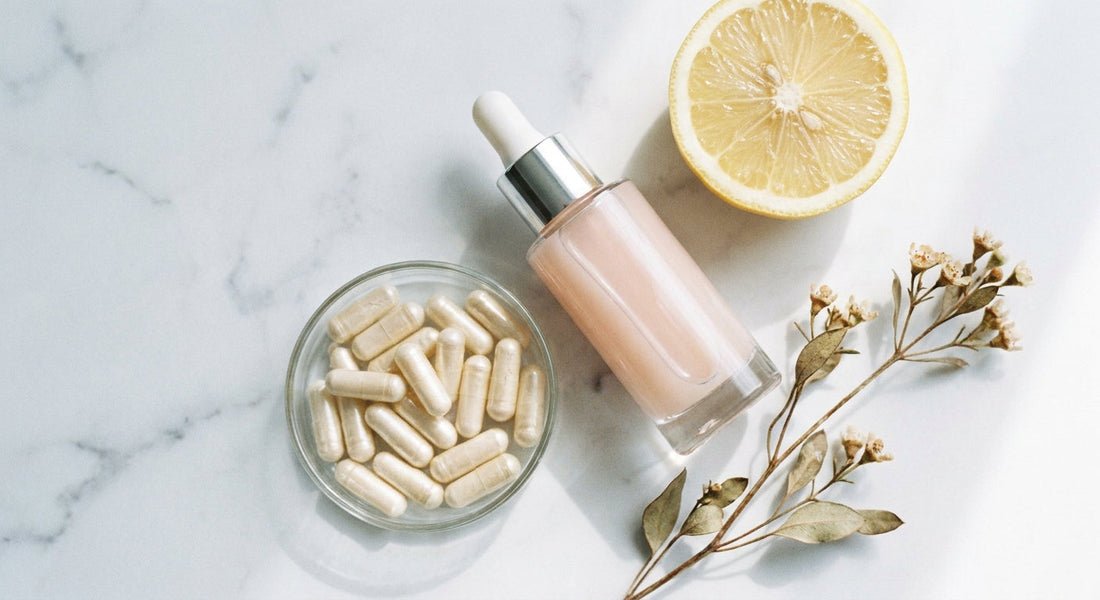 A minimalist marble flat-lay of a glass serum dropper bottle alongside supplement capsules, a sliced citrus fruit, and botanical sprigs in soft natural light.