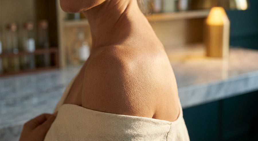 A close-up editorial photograph of a woman's shoulder and neck bathed in warm golden side light against a cool marble and amber background, evoking the science and beauty of skin aging.